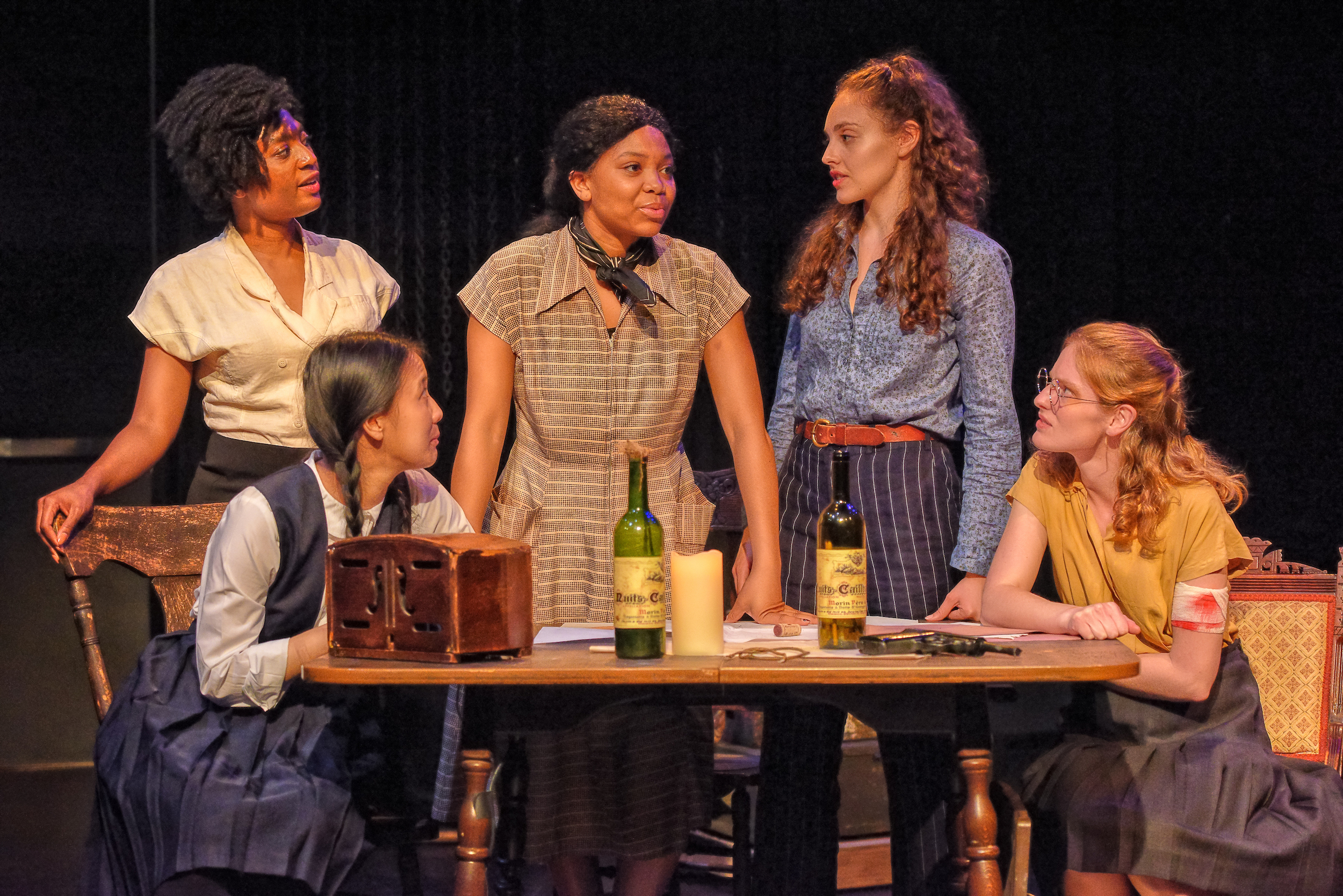 An interracial group of women stand around a table in discussion. They are dressed in period clothing from 20th century America.